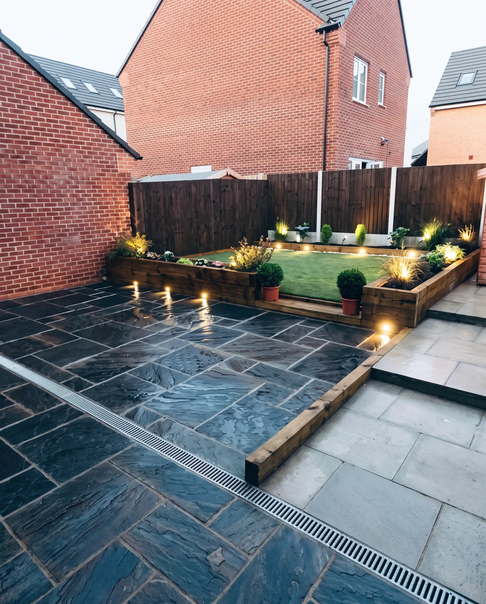 Black limestone patio with sleeper raised beds, lawn and integrated lighting at dusk, bespoke landscaping by DVH Innovations in Greater Manchester